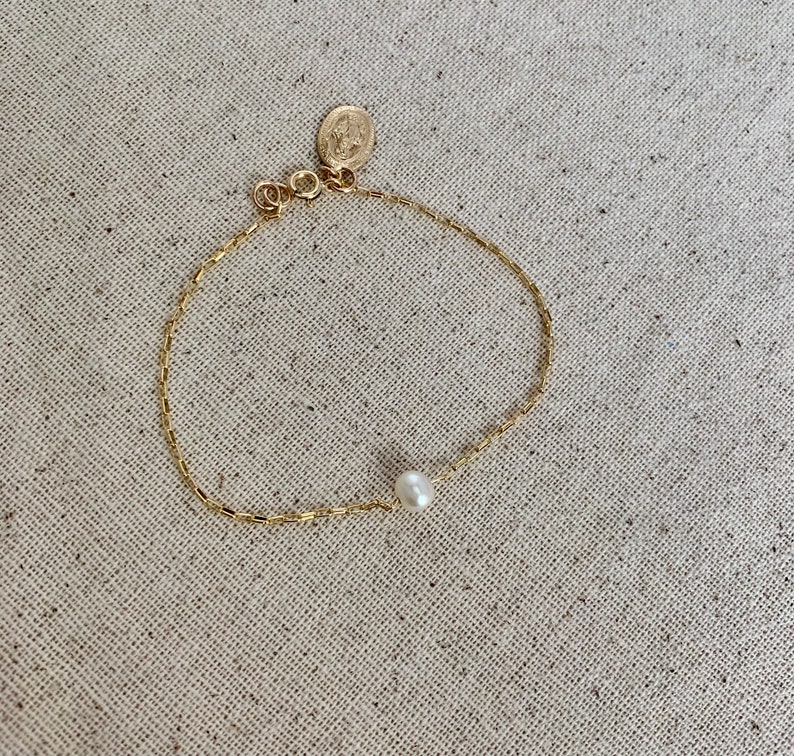 May include: Delicate gold-toned bracelet featuring a single white pearl and a small circular charm. The chain has a rectangular link design. The bracelet is displayed on a neutral-toned fabric surface.