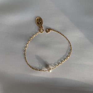 May include: A delicate gold-colored bracelet with a small pearl and a religious pendant. The bracelet features a chain-like design and a clasp. The pendant has an oval shape with a detailed relief. The bracelet is displayed on a white surface.