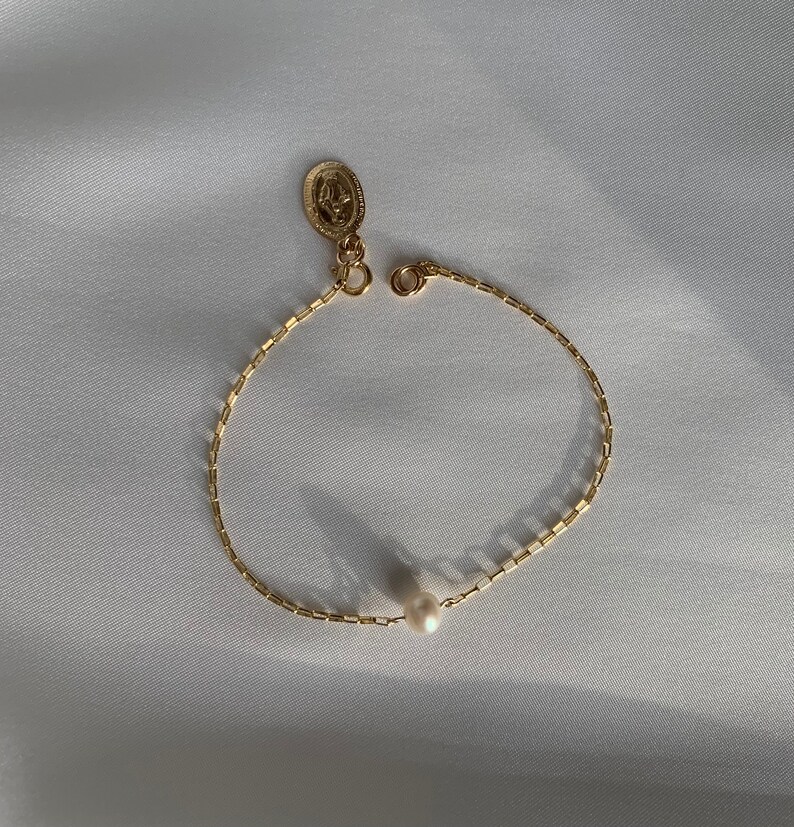 May include: A delicate gold-colored bracelet with a small pearl and a religious pendant. The bracelet features a chain-like design and a clasp. The pendant has an oval shape with a detailed relief. The bracelet is displayed on a white surface.