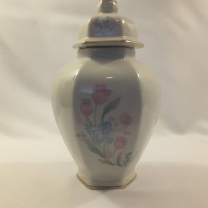 May include: A white porcelain vase with a floral design. The vase has a gold trim around the top and bottom. The flowers are pink, blue, and green.