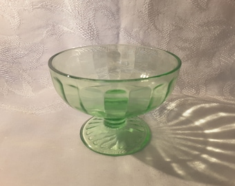 Puede incluir: Un cuenco de postre de vidrio verde vintage con base de pedestal. El cuenco tiene un diseño acanalado alrededor de los lados y un patrón decorativo en la base. El vidrio es de color verde claro y translúcido.