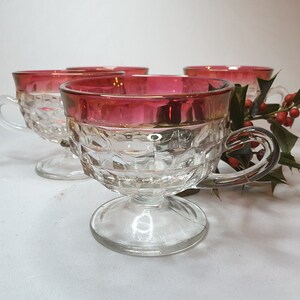 May include: Set of four clear glass teacups with a ruby red rim and handle. The cups have a diamond pattern and sit on a pedestal base. A sprig of holly with red berries is in the background.