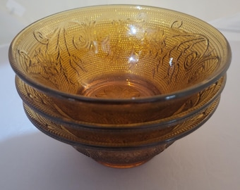 Pode incluir: Três tigelas de vidro âmbar empilhadas. Cada tigela tem uma superfície texturizada e um desenho floral na borda. As tigelas estão vazias e parecem ser vintage. A cor é dourada.