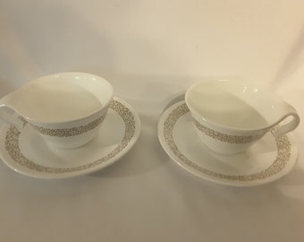 May include: Two white teacups with a brown floral pattern around the rim, each sitting on a matching saucer.