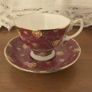 May include: A red teacup and saucer with a floral pattern of pink and yellow roses. The cup and saucer have a gold trim.
