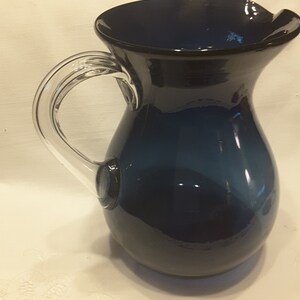 May include: A dark blue glass pitcher with a clear glass handle. The pitcher has a rounded body and a wide mouth.