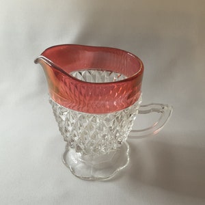 May include: A clear glass creamer with a diamond pattern and a red rim. The creamer has a small handle and a scalloped base.