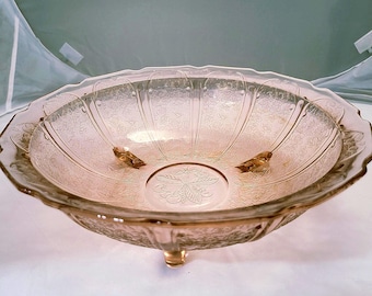 Pode incluir: Uma tigela de vidro rosa vintage com borda ondulada e três pés pequenos. A tigela apresenta um padrão floral em relevo no interior e exterior. A tigela é provavelmente usada para servir ou exibir.