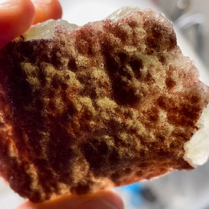 May include: A close-up of a light brown and white crystal with a textured surface. The crystal has a translucent appearance with a hint of red.