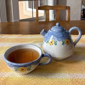 Op de afbeelding: Een blauwe en witte theepot met gele bloemenaccenten en een bijpassend theekopje gevuld met bruine thee. De theepot en het theekopje staan op een geel en wit gestreept placemat.