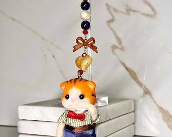Könnte beinhalten: Eine kleine orange-weiße Katzenfigur, die ein grün-weiß gestreiftes Hemd, einen braunen Gürtel und eine blaue Hose trägt. Die Katze hält eine rote Fliege in ihrer Pfote. Die Figur ist an einer goldenen Kette mit blauen, weißen und roten Perlen, einem goldenen Herz-Anhänger und einer braunen Schleife befestigt.