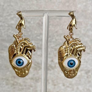 May include: Gold heart-shaped earrings with a blue eye in the center. Each earring features a small gold hand dangling from the top.