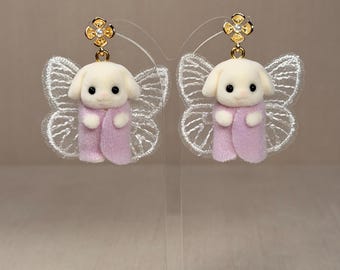 Könnte beinhalten: Ein Paar skurrile Ohrringe mit kleinen, weißen Hasenfiguren, die rosa Umhänge tragen und an zarten, spitzenbesetzten Schmetterlingsflügeln befestigt sind. Die Ohrringe haben goldfarbene Blumenstecker. Die Ohrringe werden auf einem transparenten, abgewinkelten Ständer ausgestellt.