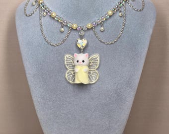 Könnte beinhalten: Eine skurrile Halskette mit einer weißen Katzenfigur mit gelben Feenflügeln und einem passenden Kleid. Die Halskette hat einen Perlenstrang mit schillernden Perlen, kleinen hängenden Perlen und silbernen Kettenakzenten. Der Anhänger enthält einen herzförmigen Charm.