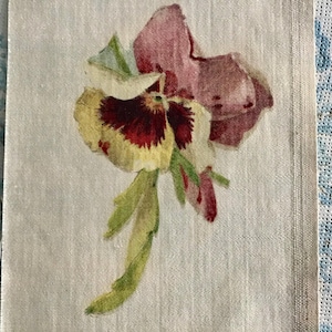 May include: A watercolor painting of a pansy flower with burgundy, yellow, and white petals, and green leaves. The flower is on a light beige fabric background.