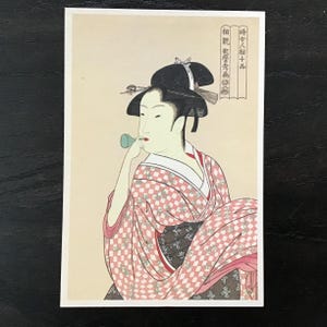 May include: A Japanese woodblock print depicting a woman in a pink and white checkered kimono blowing into a green pipe. The text in the image reads "婦女相撲 十番".