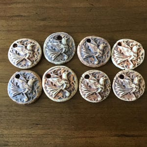 May include: Eight ceramic pendants with a bird design. Each pendant is round with a hole at the top and features a detailed relief carving of a bird surrounded by leaves. The color palette includes shades of brown, beige, and gray.