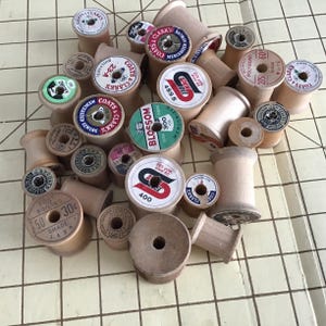 May include: A collection of vintage wooden sewing thread spools in various sizes, featuring paper labels with brand names like Coats & Clark, Spun Dee, and Blossom. The spools display printed text including yardage, shade numbers, and prices.