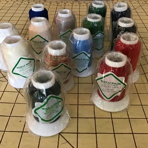 May include: A collection of fourteen spools of Woolly Nylon sewing thread, each individually wrapped in clear plastic. The spools feature white bases and tops, with labels reading Woolly Nylon, and come in various colors including red, blue, green, black, white, and beige.