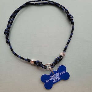 May include: A blue and black rope dog collar with a blue bone-shaped tag. The tag has white text that reads "Thurston, Sandy, 106 Winchester Road, SG19 1DP". The collar has silver-colored paw print charms.