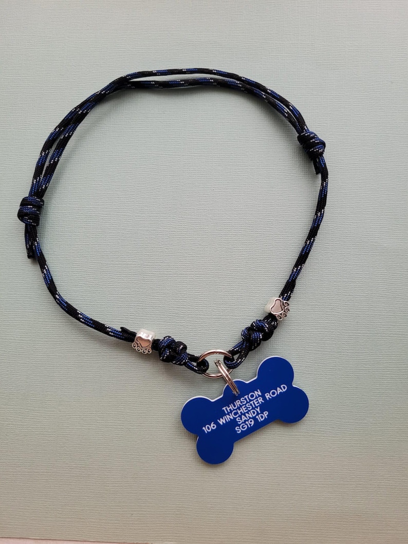 May include: A blue and black rope dog collar with a blue bone-shaped tag. The tag has white text that reads "Thurston, Sandy, 106 Winchester Road, SG19 1DP". The collar has silver-colored paw print charms.