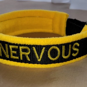May include: Black and yellow dog collar with the word "NERVOUS" embroidered in yellow thread. The collar has a soft yellow interior and a black exterior with the embroidered text. The collar is designed for a dog.