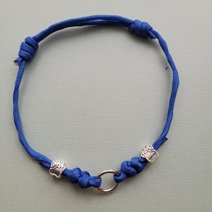 May include: A blue cord bracelet with a silver ring and two silver paw print charms. The bracelet is knotted and designed for pets. The cord is a vibrant blue color, and the charms are detailed.