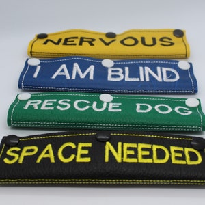 May include: Four different dog leash attachments with velcro closures. The attachments are made of leather and have different coloured backgrounds. The text on the attachments reads "NERVOUS", "I AM BLIND", "RESCUE DOG", and "SPACE NEEDED".