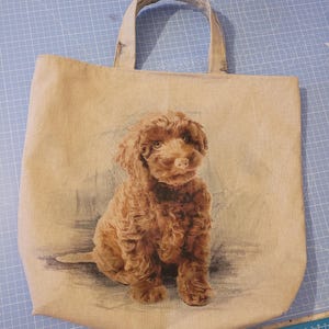 May include: A brown canvas tote bag with a printed image of a brown puppy dog. The dog is sitting and looking at the camera.