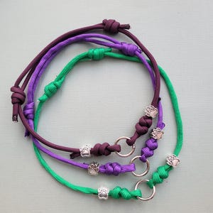 May include: Three handmade dog collars in purple, green, and burgundy. Each collar features a silver paw print charm and a metal ring. The collars are made of knotted cord.