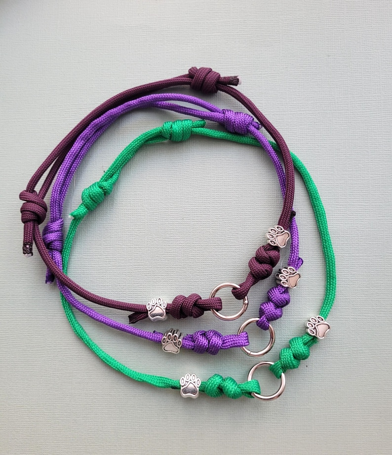 May include: Three handmade dog collars in purple, green, and burgundy. Each collar features a silver paw print charm and a metal ring. The collars are made of knotted cord.