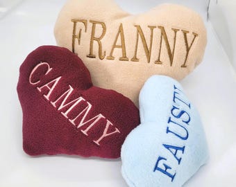 Puede incluir: Tres cojines de felpa en forma de corazón en crema, burdeos y azul claro. Cada cojín tiene un nombre bordado: "FRANNY" en dorado, "CAMMY" en blanco y "FAUST" en azul. Los cojines son suaves y esponjosos.
