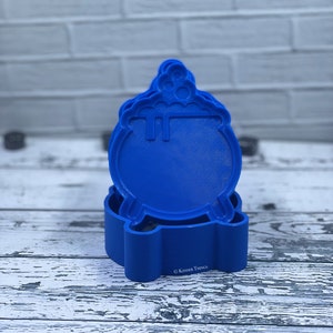 May include: Blue plastic cookie cutter shaped like a cauldron with a handle and a base. The cutter is stacked on top of itself. The text "Kinder Things" is visible on the base of the cutter.