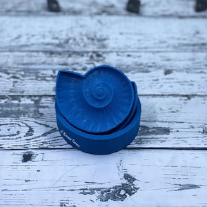 May include: A blue silicone mold shaped like a nautilus shell. The mold is in two pieces, with the top piece showing the spiral of the shell. The mold is on a white wooden surface.