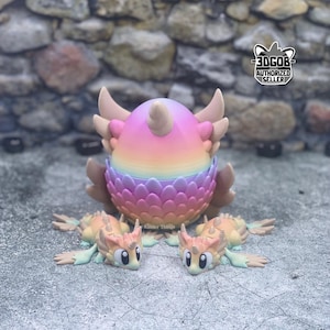 May include: A colorful, 3D-printed fantasy creature egg with a horn, wings, and a layered base. Two small, matching creatures are positioned in front. The egg is a gradient of pink, yellow, and blue. The background is a stone wall.
