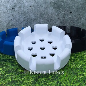 May include: Three round, decorative holders in blue, white, and black. The white holder is in the foreground, featuring heart-shaped cutouts. Each holder has a series of curved, raised sections around the perimeter. The text "Kinder Things" is visible.