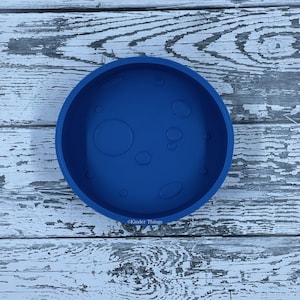 May include: A round, deep blue silicone bowl with a moon-like design. The bowl features various circular and oval indentations, resembling craters. The text "Kinder Things" is printed on the side of the bowl.