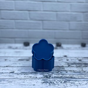 May include: A set of three blue silicone flower-shaped molds. The molds are stacked on top of each other. The molds are on a white wooden surface. © Kinder Things