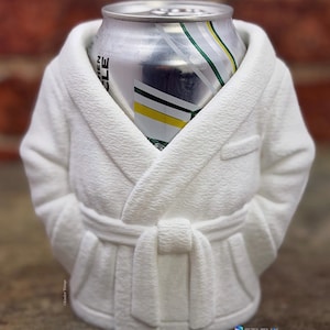 May include: A white, textured can cooler shaped like a bathrobe, with a silver can inside. The bathrobe has a tie and lapels, and the can has green and yellow stripes. The cooler is designed to keep beverages cold.