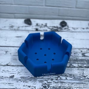 May include: A blue, hexagonal ashtray with multiple grooves for resting items. The ashtray has several small holes in the center and the text "© Kinder Things" is printed on the side. The ashtray is made of a flexible material.
