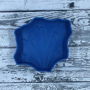 May include: Blue silicone mold shaped like a firework with stars. The mold is on a white wooden surface.