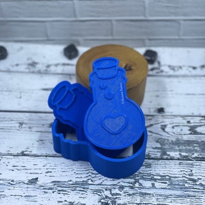 May include: Blue plastic cookie cutters shaped like a potion bottle with a heart and a smaller bottle. The cutters are on a white wooden surface.