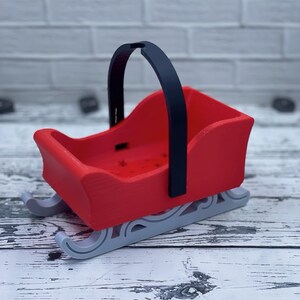 May include: A red and black miniature sleigh with a black handle. The sleigh has a white base and is made of plastic.