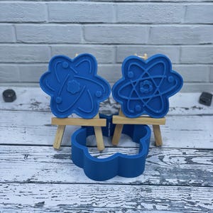 May include: Three blue silicone cookie cutters, two shaped like atoms and one shaped like a flower, displayed on small wooden easels.