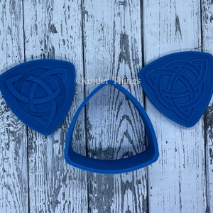 May include: Three blue silicone cookie cutters shaped like triangles with a Celtic knot design. The cutters are on a white wooden background.