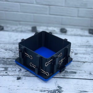 May include: A black plastic box with a blue interior, held together with binder clips. The box is sitting on a white wooden surface.