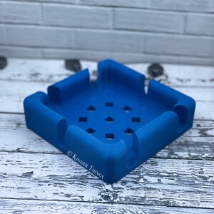 May include: A blue silicone phone stand with a square base and a raised edge. The stand has a grid of holes on the bottom and the text "© KINDER THINGS" is printed on the side.