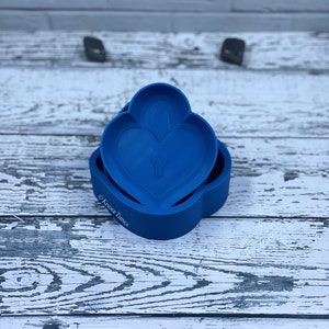 Puede incluir: Un molde de plástico azul con forma de corazón con un corazón más pequeño en su interior. El molde está sobre una superficie de madera blanca.