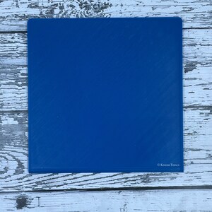 May include: A solid blue square cutting mat with a textured surface. The mat has the text "Kinder Things" in white lettering in the bottom right corner.