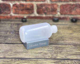 May include: A clear plastic bottle with a screw-on cap, resting on a gray block with the text "@Kinder Things" printed on it.